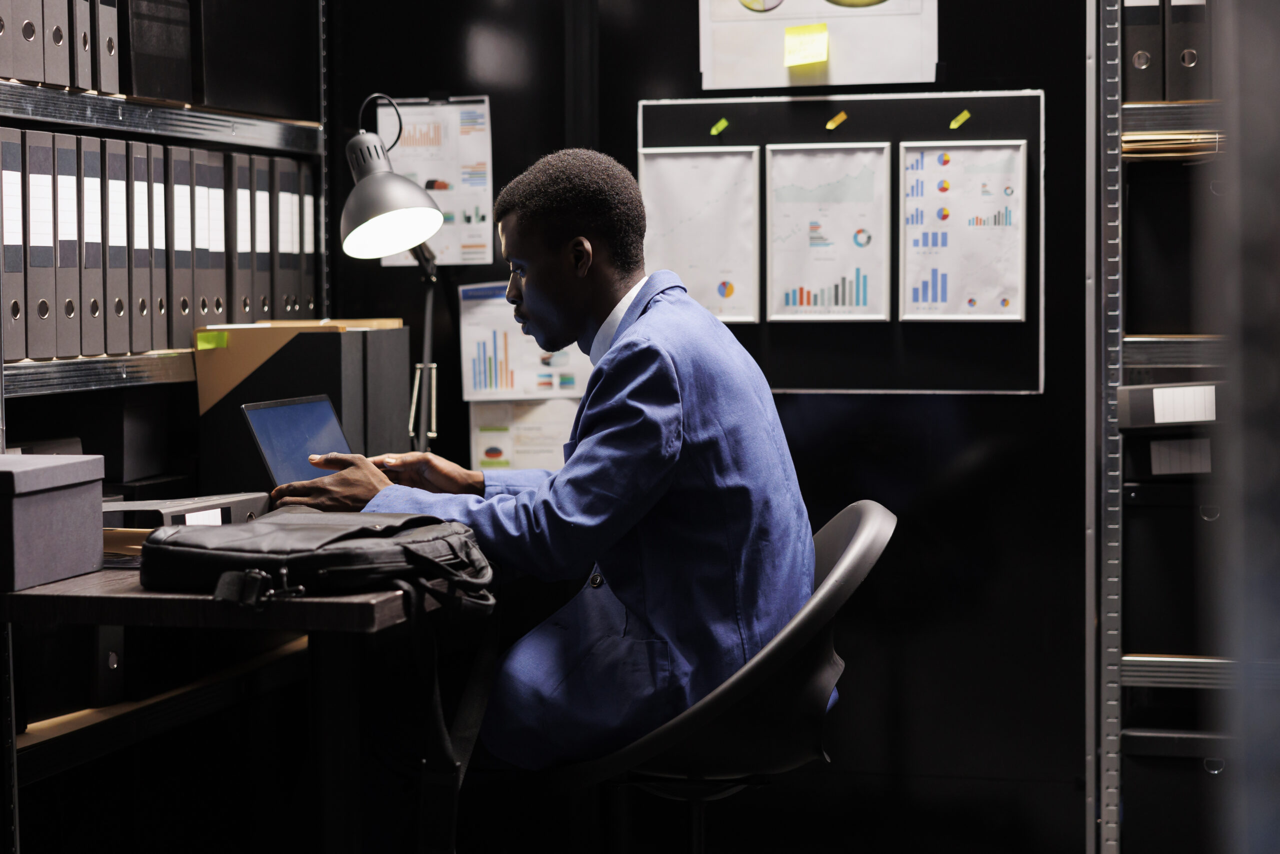 Bookkeeper reading administrative report, organizing management documents in storage room. African american businessman wearing formal suit working late at night in corporate depository